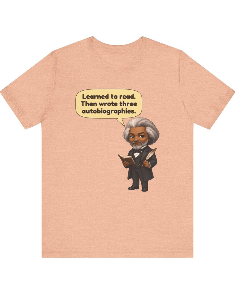 Puede incluir: Una camiseta de color melocot&oacute;n con una ilustraci&oacute;n de dibujos animados de Frederick Douglass sosteniendo un libro y una pluma. Un globo de texto sobre la figura contiene el texto: Learned to read. Then wrote three autobiographies.
