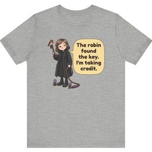 May include: A heather gray t-shirt featuring a cartoon illustration of a person in a black coat holding a large key with a robin perched on it. A speech bubble contains the text: The robin found the key. I'm taking credit. Small purple flowers are at the feet.