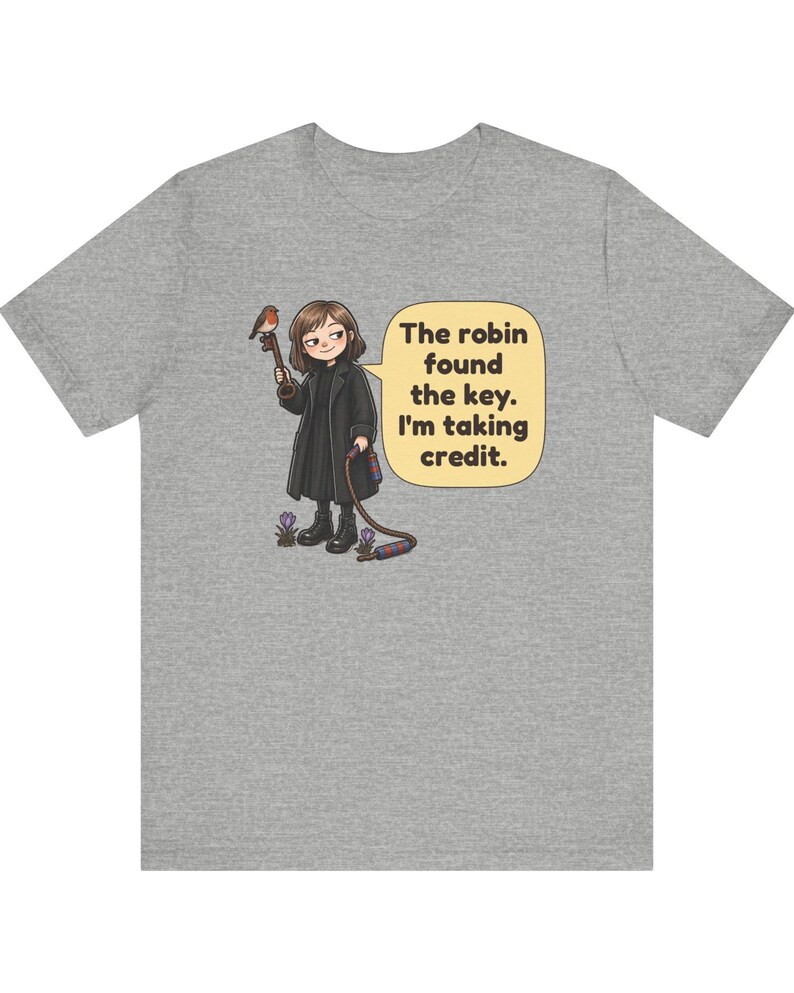 May include: A heather gray t-shirt featuring a cartoon illustration of a person in a black coat holding a large key with a robin perched on it. A speech bubble contains the text: The robin found the key. I'm taking credit. Small purple flowers are at the feet.