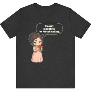 K&ouml;nnte beinhalten: Ein dunkelgraues T-Shirt mit einer Cartoon-Illustration einer Person mit braunen Haaren in einem hellrosa Kleid, die zwei rote Herzen h&auml;lt. Eine Sprechblase &uuml;ber der Figur enth&auml;lt den Text: I'm not meddling. I'm matchmaking.