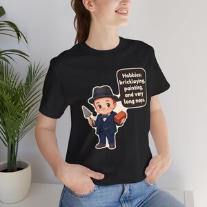 Pu&ograve; includere: Una t-shirt nera con un'illustrazione a cartone animato di una persona in abito scuro e fedora che tiene in mano una cazzuola e un mattone. Un fumetto contiene il testo: Hobbies: bricklaying, painting, and very long naps.