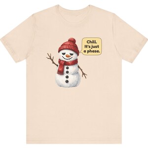 Pode incluir: Uma t-shirt de manga curta em bege claro com um gr&aacute;fico de um boneco de neve a usar um gorro e um cachecol de malha vermelhos. O boneco de neve tem uma express&atilde;o c&eacute;tica e acena. Um bal&atilde;o de fala ao lado cont&eacute;m o texto: Chill. It's just a phase.