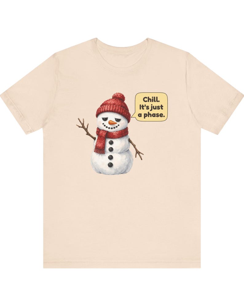Pode incluir: Uma t-shirt de manga curta em bege claro com um gr&aacute;fico de um boneco de neve a usar um gorro e um cachecol de malha vermelhos. O boneco de neve tem uma express&atilde;o c&eacute;tica e acena. Um bal&atilde;o de fala ao lado cont&eacute;m o texto: Chill. It's just a phase.