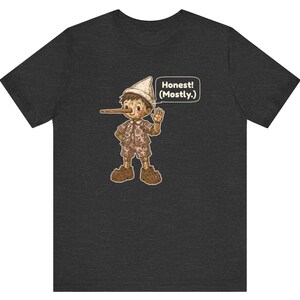 K&ouml;nnte beinhalten: Ein dunkelgraues Kurzarm-T-Shirt mit einer Grafik einer Holzpuppe mit langer Nase, die einen Hut und ein gemustertes Outfit tr&auml;gt. Eine Sprechblase neben der Figur enth&auml;lt den Text, Honest! (Mostly.)