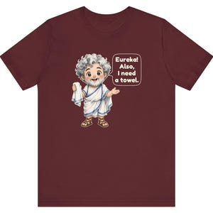 May include: A maroon short-sleeve t-shirt featuring a cartoon illustration of a bearded man in a white toga holding a towel. A speech bubble next to the character contains the text: Eureka! Also, I need a towel. The design is centered on the chest.