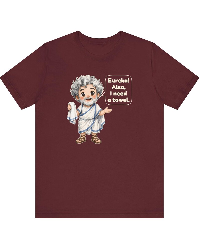 May include: A maroon short-sleeve t-shirt featuring a cartoon illustration of a bearded man in a white toga holding a towel. A speech bubble next to the character contains the text: Eureka! Also, I need a towel. The design is centered on the chest.