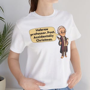 Pu&ograve; includere: Una t-shirt bianca a maniche corte con un'illustrazione a cartone animato di un uomo con gli occhiali che tiene in mano una penna e una pergamena. Un fumetto giallo contiene il testo: Hebrew professor. Poet. Accidentally Christmas.