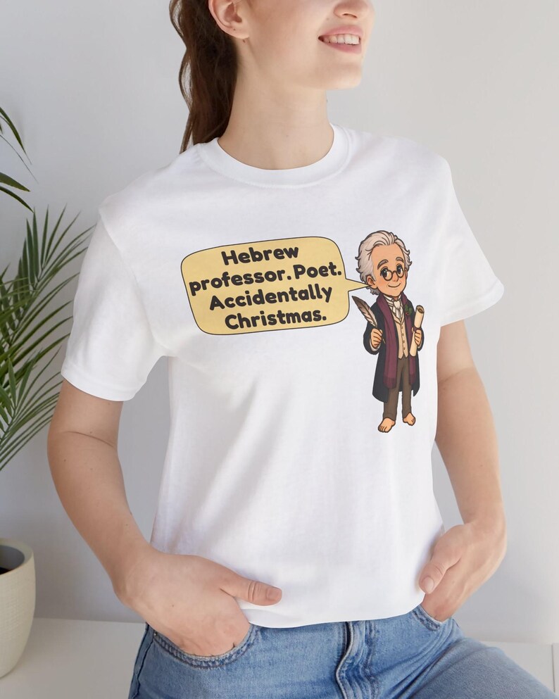 Pu&ograve; includere: Una t-shirt bianca a maniche corte con un'illustrazione a cartone animato di un uomo con gli occhiali che tiene in mano una penna e una pergamena. Un fumetto giallo contiene il testo: Hebrew professor. Poet. Accidentally Christmas.