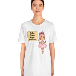May include: A white t-shirt featuring a cartoon illustration of a character in a pink tutu and tiara holding a star-tipped wand. A yellow speech bubble next to the character contains the text: Tutu. Wand. Sugar addiction.