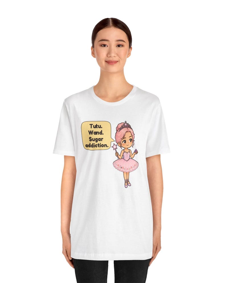 May include: A white t-shirt featuring a cartoon illustration of a character in a pink tutu and tiara holding a star-tipped wand. A yellow speech bubble next to the character contains the text: Tutu. Wand. Sugar addiction.