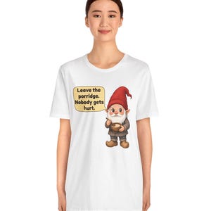 May include: A white t-shirt featuring a graphic of a gnome wearing a red hat and brown clothing, holding a bowl of porridge. A yellow speech bubble next to the gnome contains the text: Leave the porridge. Nobody gets hurt.