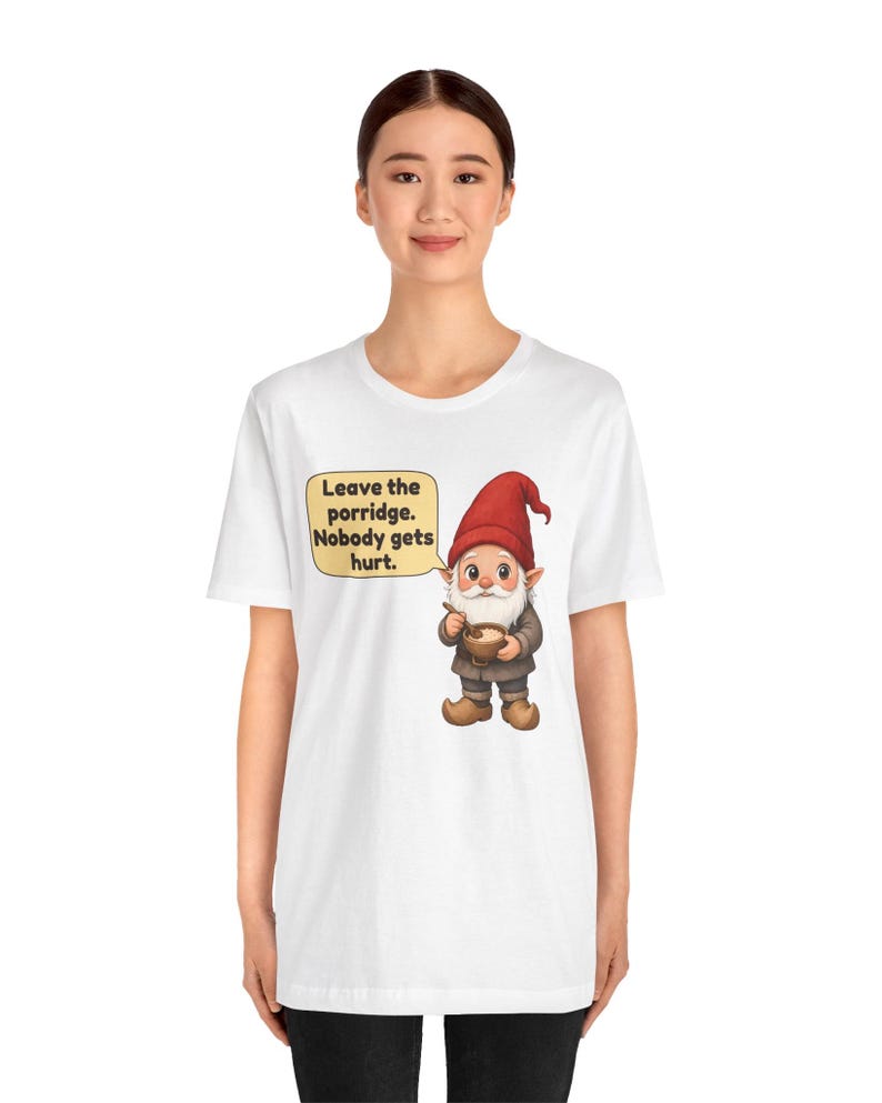 May include: A white t-shirt featuring a graphic of a gnome wearing a red hat and brown clothing, holding a bowl of porridge. A yellow speech bubble next to the gnome contains the text: Leave the porridge. Nobody gets hurt.
