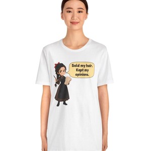 May include: A white short-sleeve t-shirt featuring a cartoon illustration of a person with dark braided hair, a red bow, and a dark dress. A speech bubble next to the character contains the text: Sold my hair. Kept my opinions.
