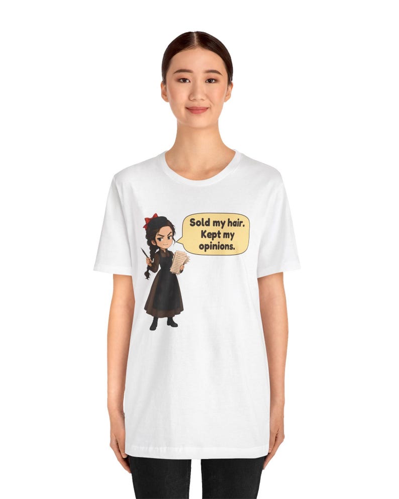 May include: A white short-sleeve t-shirt featuring a cartoon illustration of a person with dark braided hair, a red bow, and a dark dress. A speech bubble next to the character contains the text: Sold my hair. Kept my opinions.