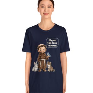 May include: A navy blue t-shirt featuring a graphic of a monk in a brown robe standing with a white rabbit, a brown dog, and a blue bird. A speech bubble above the monk contains the text: My pets talk to me. Yours too?
