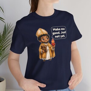 May include: A navy blue t-shirt featuring a cartoon illustration of a bishop holding a book and a flaming heart. A speech bubble next to the figure contains the text: Make me good. Just not yet. The shirt has short sleeves and a crew neckline.