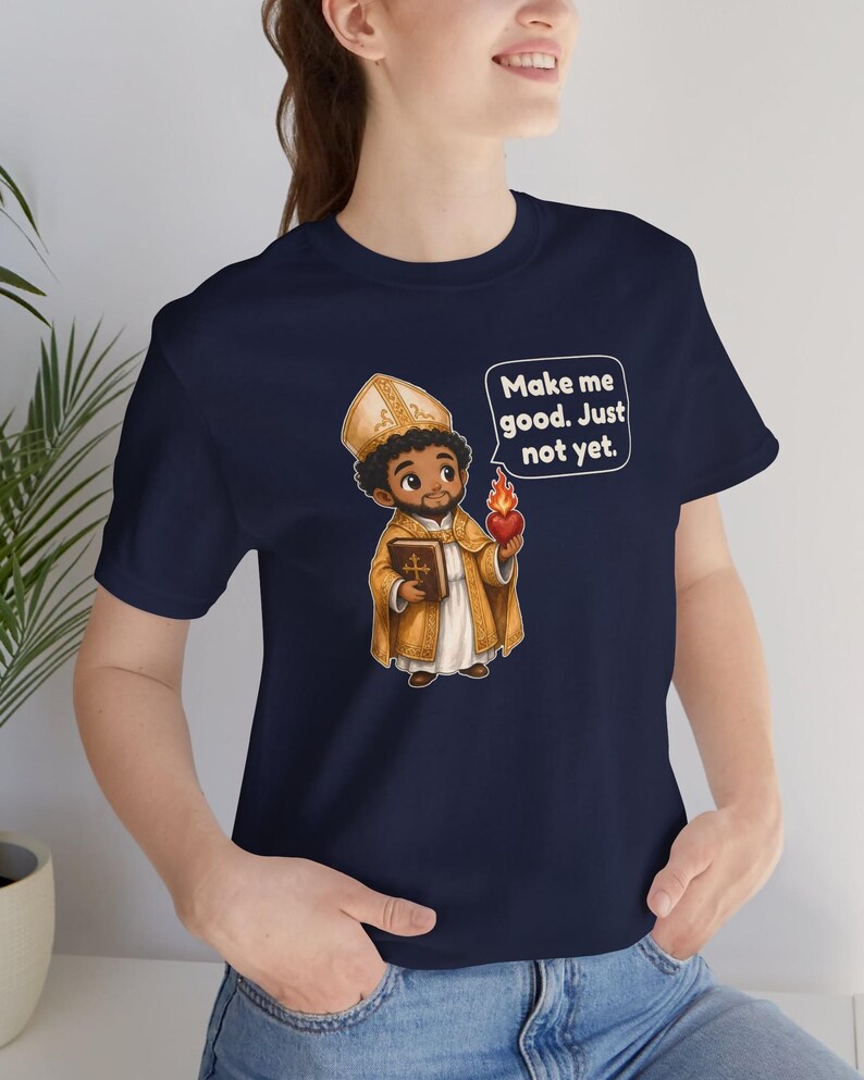 May include: A navy blue t-shirt featuring a cartoon illustration of a bishop holding a book and a flaming heart. A speech bubble next to the figure contains the text: Make me good. Just not yet. The shirt has short sleeves and a crew neckline.