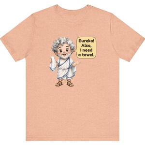 May include: A peach-colored t-shirt featuring a cartoon illustration of a bearded man in a white toga and brown sandals holding a white towel. A speech bubble next to the figure contains the text: Eureka! Also, I need a towel.