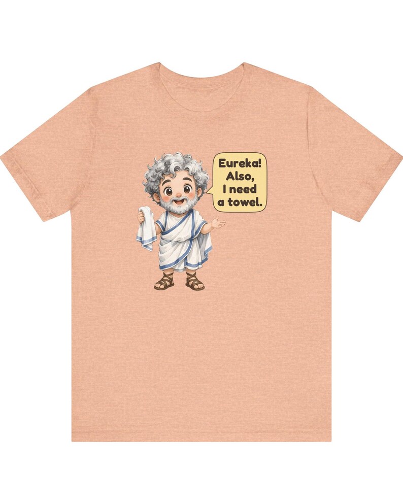 May include: A peach-colored t-shirt featuring a cartoon illustration of a bearded man in a white toga and brown sandals holding a white towel. A speech bubble next to the figure contains the text: Eureka! Also, I need a towel.