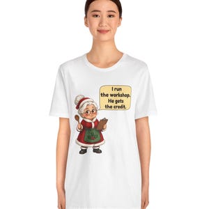 K&ouml;nnte beinhalten: Ein wei&szlig;es T-Shirt mit einer Grafik einer &auml;lteren Frau in einem rot-wei&szlig;en Festtagsoutfit mit gr&uuml;ner Sch&uuml;rze. Sie h&auml;lt einen Holzl&ouml;ffel und ein Klemmbrett. Eine Sprechblase enth&auml;lt den Text: I run the workshop. He gets the credit.