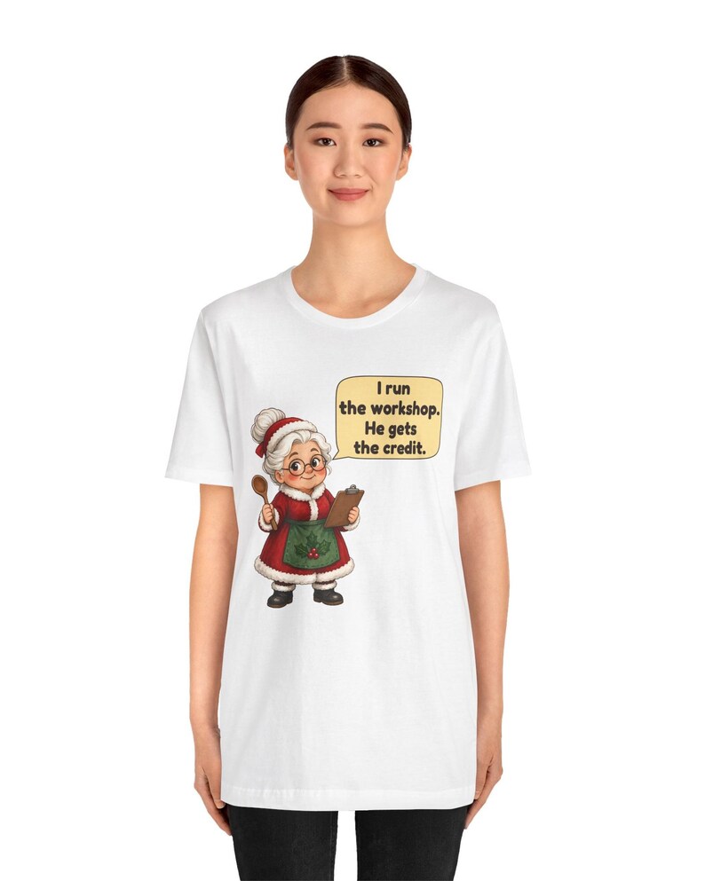 K&ouml;nnte beinhalten: Ein wei&szlig;es T-Shirt mit einer Grafik einer &auml;lteren Frau in einem rot-wei&szlig;en Festtagsoutfit mit gr&uuml;ner Sch&uuml;rze. Sie h&auml;lt einen Holzl&ouml;ffel und ein Klemmbrett. Eine Sprechblase enth&auml;lt den Text: I run the workshop. He gets the credit.