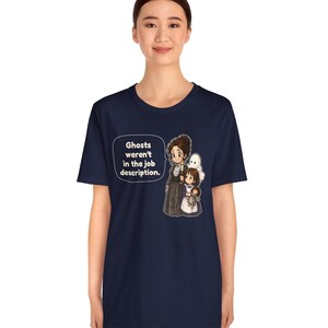 Op de afbeelding: Een marineblauw t-shirt met een grafische illustratie van een vrouw en een kind dat een pop vasthoudt, vergezeld door een witte geestfiguur. Een tekstballon naast de figuren bevat de tekst: Ghosts weren't in the job description.