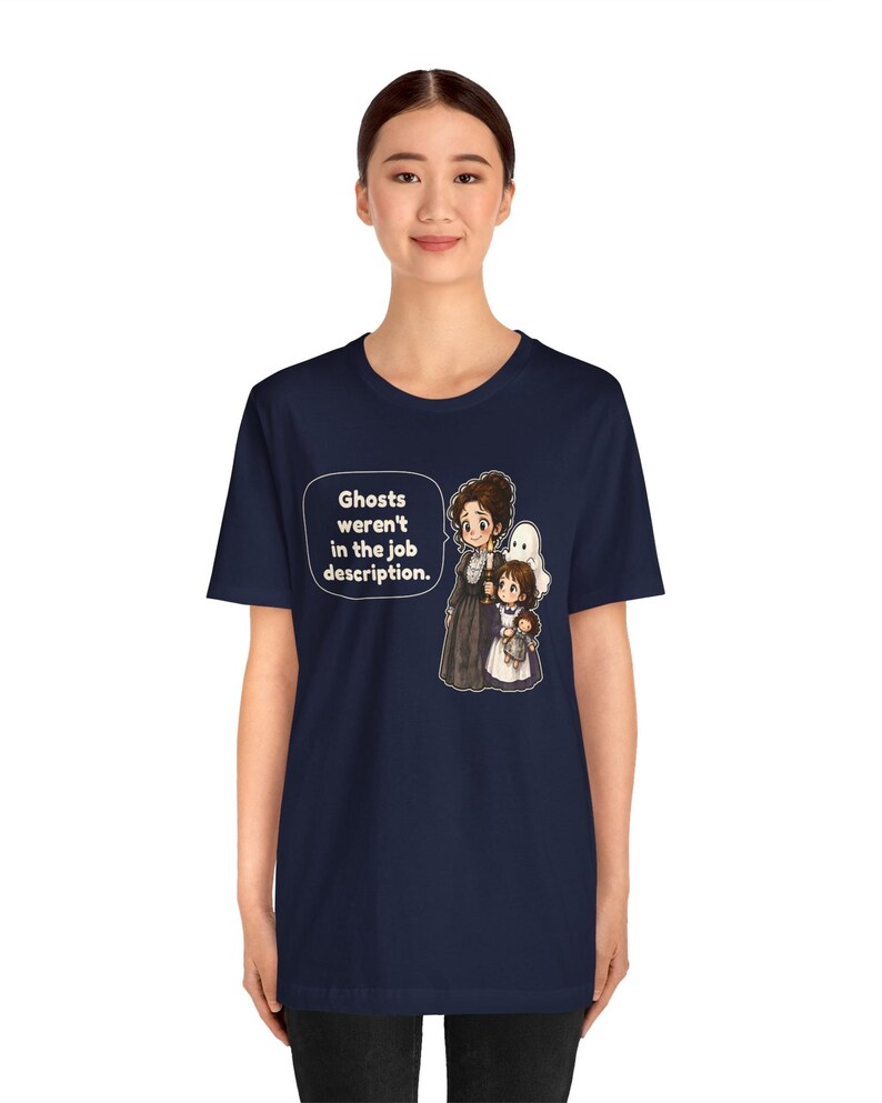 Op de afbeelding: Een marineblauw t-shirt met een grafische illustratie van een vrouw en een kind dat een pop vasthoudt, vergezeld door een witte geestfiguur. Een tekstballon naast de figuren bevat de tekst: Ghosts weren't in the job description.