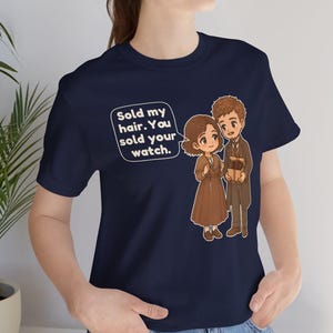 May include: A navy blue t-shirt featuring a cartoon illustration of two figures and a speech bubble with the text: Sold my hair. You sold your watch. The figures are dressed in brown and tan clothing, with one holding a small gift box.