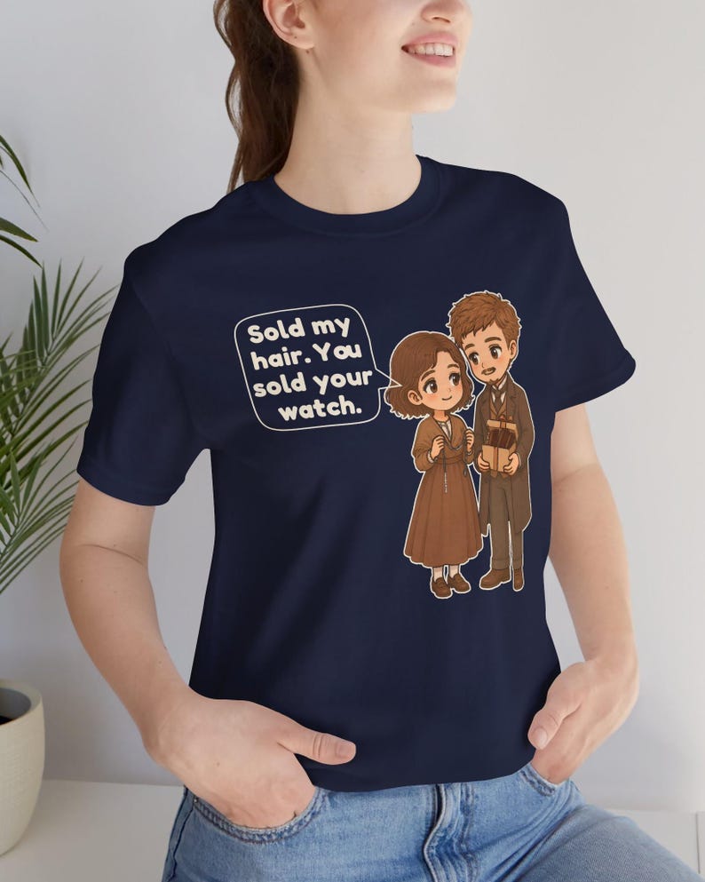 May include: A navy blue t-shirt featuring a cartoon illustration of two figures and a speech bubble with the text: Sold my hair. You sold your watch. The figures are dressed in brown and tan clothing, with one holding a small gift box.