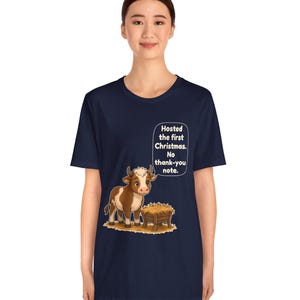 以下が含まれることがあります： 紺色のTシャツで、干し草が入った木製の飼い葉桶の横に立つ茶色と白の牛のグラフィックが描かれています。牛の上にある吹き出しには、白い文字で「Hosted the first Christmas. No thank-you note.」と書かれています。