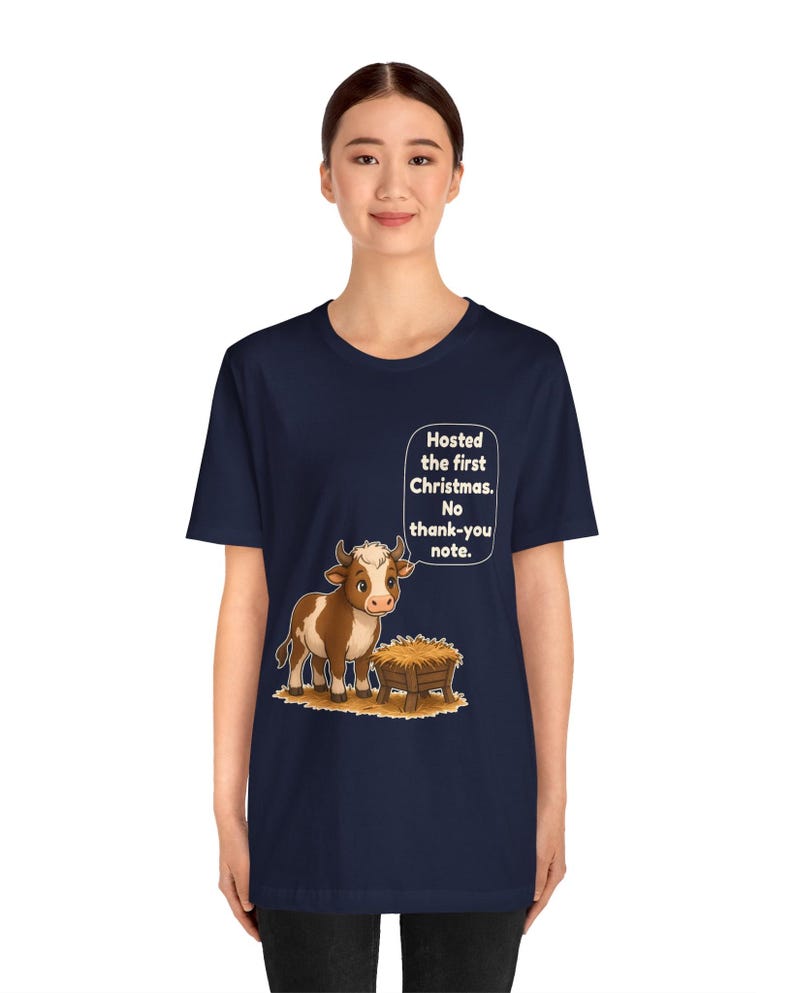 以下が含まれることがあります： 紺色のTシャツで、干し草が入った木製の飼い葉桶の横に立つ茶色と白の牛のグラフィックが描かれています。牛の上にある吹き出しには、白い文字で「Hosted the first Christmas. No thank-you note.」と書かれています。