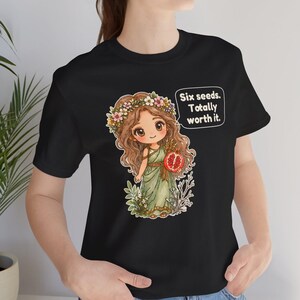May include: A black t-shirt featuring a graphic of a cartoon character with long brown hair, a flower crown, and a green dress, holding a pomegranate. A speech bubble next to the character contains the text: Six seeds. Totally worth it.