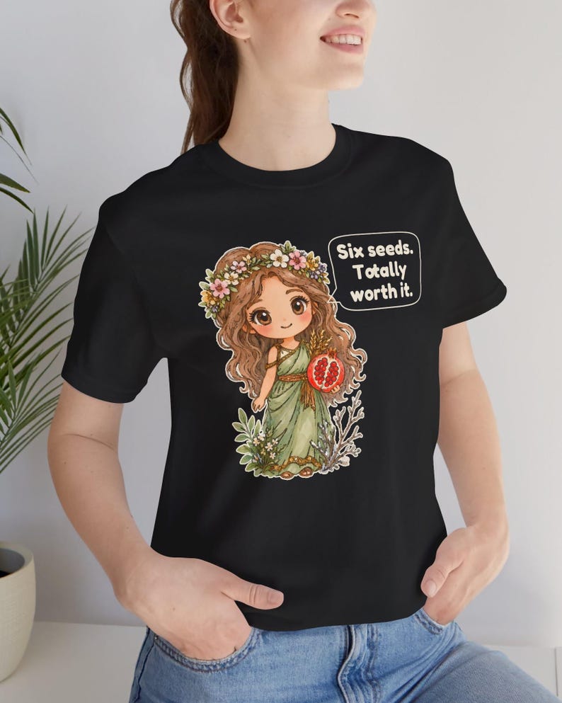 May include: A black t-shirt featuring a graphic of a cartoon character with long brown hair, a flower crown, and a green dress, holding a pomegranate. A speech bubble next to the character contains the text: Six seeds. Totally worth it.