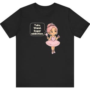 May include: A black t-shirt featuring a cartoon illustration of a ballerina with pink hair, a pink tutu, and a tiara. The character holds a star-tipped wand and a cupcake. A speech bubble contains the text: Tutu. Wand. Sugar addiction.