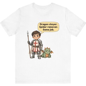 K&ouml;nnte beinhalten: Ein wei&szlig;es T-Shirt mit einer Grafik eines Cartoon-Ritters in silberner R&uuml;stung, der einen Speer h&auml;lt und neben einem kleinen gr&uuml;nen Drachen steht. Eine Sprechblase &uuml;ber dem Ritter enth&auml;lt den Text: Dragon slayer. Spider remover. Same job.