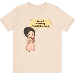 K&ouml;nnte beinhalten: Ein hellbeiges T-Shirt mit einer Cartoon-Illustration einer Person mit dunklen lockigen Haaren, die ein hellrosa Kleid und eine Jacke tr&auml;gt und zwei rote Herzen h&auml;lt. Eine gelbe Sprechblase dar&uuml;ber enth&auml;lt den Text: I'm not meddling. I'm matchmaking.