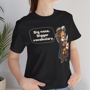 May include: A black t-shirt featuring a cartoon illustration of a historical figure with a large nose, wearing a hat with a feather and a cape. A speech bubble next to the character contains the text: Big nose. Bigger vocabulary.
