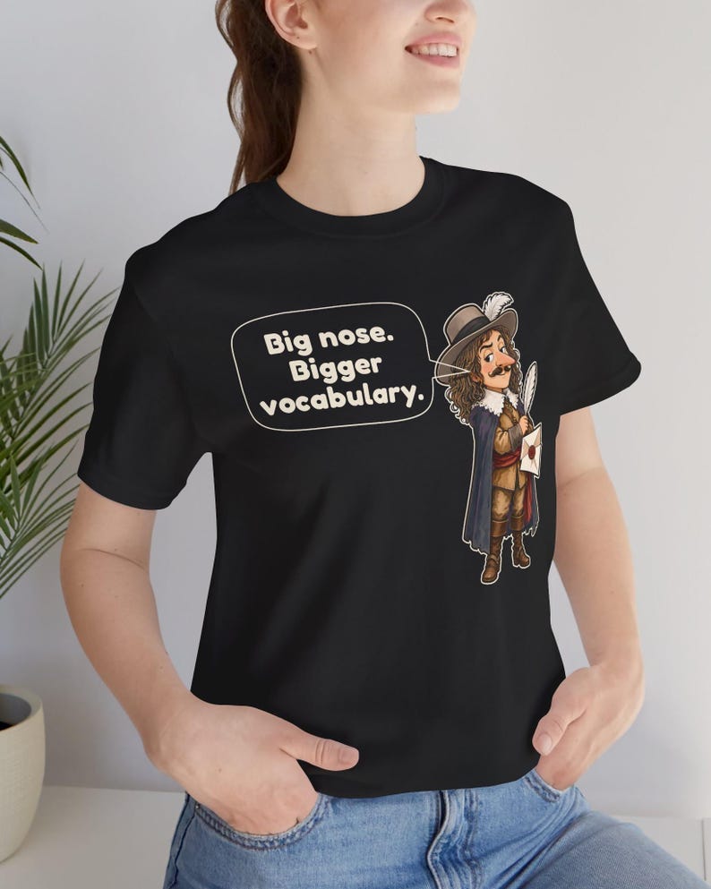 May include: A black t-shirt featuring a cartoon illustration of a historical figure with a large nose, wearing a hat with a feather and a cape. A speech bubble next to the character contains the text: Big nose. Bigger vocabulary.