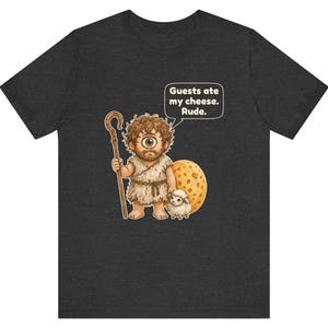 May include: A dark gray t-shirt featuring a cartoon cyclops holding a wooden staff and standing next to a small sheep and a large piece of Swiss cheese. A speech bubble above the cyclops contains the text: Guests ate my cheese. Rude.
