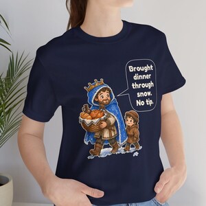 May include: A navy blue short-sleeve t-shirt featuring a graphic of a king in a blue cloak carrying a basket of food, accompanied by a child. A speech bubble contains the text: Brought dinner through snow. No tip.