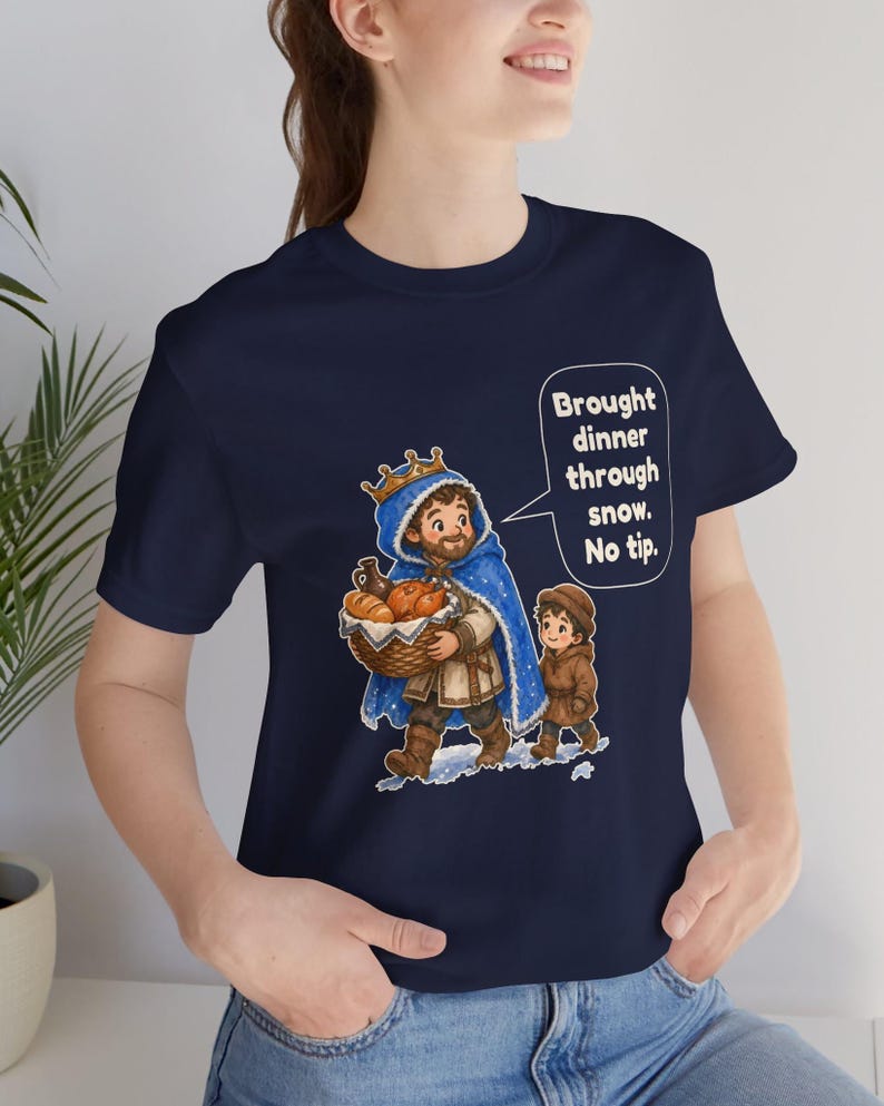 May include: A navy blue short-sleeve t-shirt featuring a graphic of a king in a blue cloak carrying a basket of food, accompanied by a child. A speech bubble contains the text: Brought dinner through snow. No tip.