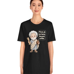 May include: A black t-shirt featuring a cartoon illustration of a bearded man in a toga holding a scroll and a brown jug. A speech bubble next to the figure contains the text: First, do no harm. Second, coffee. The design is centered on the chest.