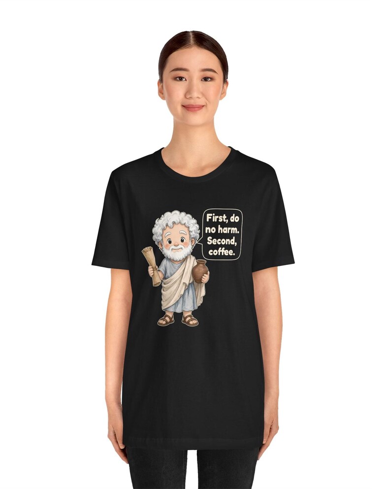 May include: A black t-shirt featuring a cartoon illustration of a bearded man in a toga holding a scroll and a brown jug. A speech bubble next to the figure contains the text: First, do no harm. Second, coffee. The design is centered on the chest.