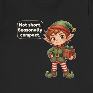 May include: A black t-shirt featuring a cartoon elf with brown hair, wearing a green tunic, red and white striped leggings, and green shoes with bells. The elf holds a red gift box and a speech bubble says, Not short. Seasonally compact.