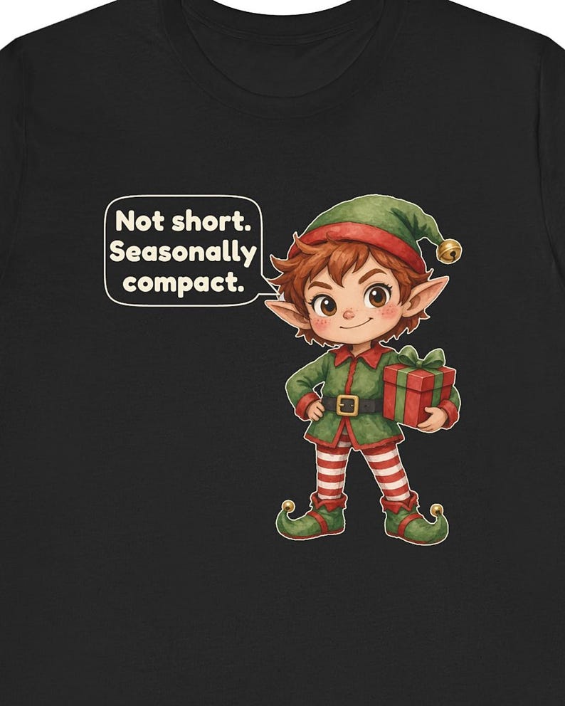 May include: A black t-shirt featuring a cartoon elf with brown hair, wearing a green tunic, red and white striped leggings, and green shoes with bells. The elf holds a red gift box and a speech bubble says, Not short. Seasonally compact.