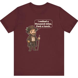 May include: A maroon short-sleeve t-shirt featuring a cartoon illustration of a bearded traveler in a brown hat and coat holding a walking stick. A speech bubble above the character contains the text: I walked a thousand miles. Pack a lunch.