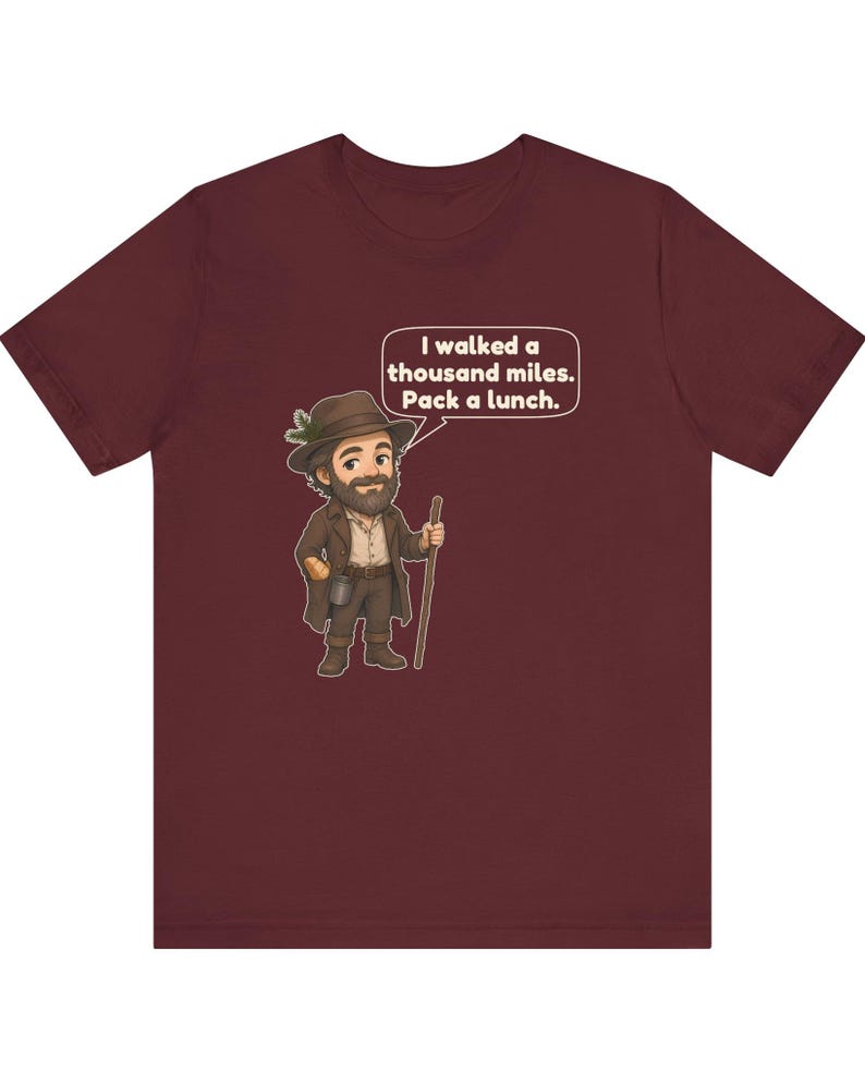 May include: A maroon short-sleeve t-shirt featuring a cartoon illustration of a bearded traveler in a brown hat and coat holding a walking stick. A speech bubble above the character contains the text: I walked a thousand miles. Pack a lunch.