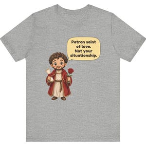 Puede incluir: Una camiseta gris jaspeado con una ilustraci&oacute;n de dibujos animados de una figura barbuda con t&uacute;nica roja que sostiene un pergamino y una rosa roja. Un globo de texto contiene: Patron saint of love. Not your situationship.