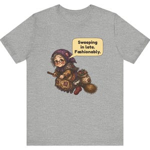 Puede incluir: Una camiseta gris jaspeado con un gr&aacute;fico de una persona mayor con gafas y pa&ntilde;uelo en la cabeza montada en una escoba. Un bocadillo sobre la figura contiene el texto: Sweeping in late. Fashionably. La ilustraci&oacute;n incluye un saco con objetos y un calcet&iacute;n rojo.