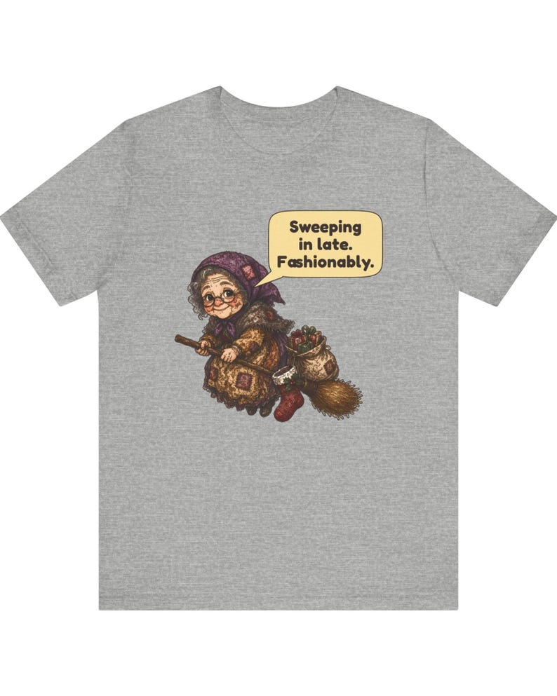 Puede incluir: Una camiseta gris jaspeado con un gr&aacute;fico de una persona mayor con gafas y pa&ntilde;uelo en la cabeza montada en una escoba. Un bocadillo sobre la figura contiene el texto: Sweeping in late. Fashionably. La ilustraci&oacute;n incluye un saco con objetos y un calcet&iacute;n rojo.